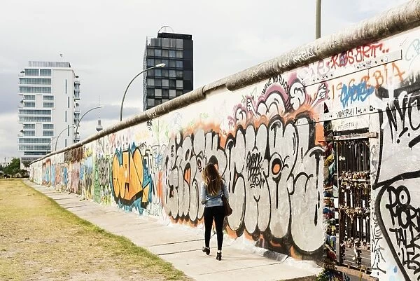 Young woman walking past graffiti on former Berlin Wall, East Side Gallery, Muhlenstrasse