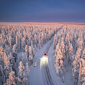 Aerial view of car on icy road and illuminated headlamps driving in the snowcapped forest, at dawn, Akaslompolo, Kolari, Pallas-Yllastunturi National Park, Lapland region, Finland, Europe