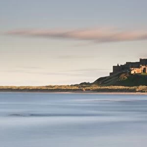 Bamburgh Castle bathed in golden evening light overlooking Bamburgh Bay with the sea filling the foreground, Northumberland, England, United