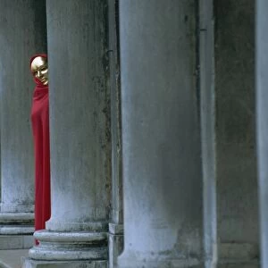 Carnival model in red cape and gold mask peering from columns in St