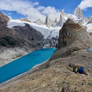 A couple in mountain gear rests on rocks with view to Lago de los Tres and Mount Fitz Roy
