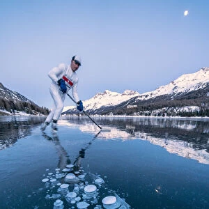 Ice hockey player man skating on Lake Sils covered in ice bubbles at dusk, Engadine
