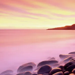 An imposing silhouette of Dunstanburgh Castle against a magnificent sky at sunrise with a beach of basalt boulders in the foreground, Embleton Bay, near Alnwick, Northumberland, England, United