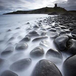 Long exposure to record motion in the sea as it washes around black basalt boulders on a beach at Embleton Bay with the silhouette of Dunstanburgh Castle in the distance, Embleton Bay, near Alnwick, Northumberland, England, United Kingdom, Europe