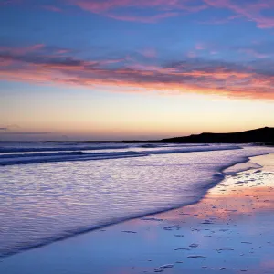 Looking across Embleton Bay at sunrise towards the silhouetted ruins of Dunstanburgh Castle in the distance and the vivid colours in the sky reflecting in the sea and wet sand, Embleton, near Alnwick, Northumberland, England, United Kingdom