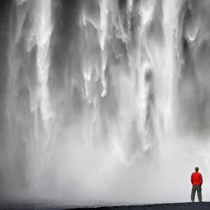 Man in red jacket standing in front of the 62m high Skogafoss waterfall near the village of Skogar