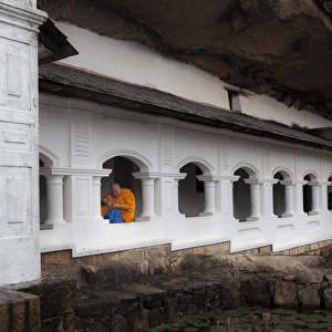 A monk sits and looks at his phone beneath shelter, Royal Rock Temple, Golden Temple of Dambulla, UNESCO World Heritage Site, Dambulla, Sri Lanka, Asia