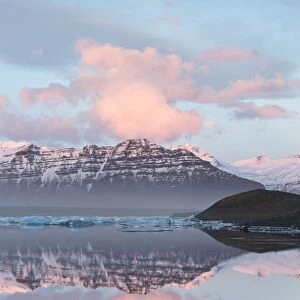 Panoramic view across the calm water of Jokulsarlon glacial lagoon towards snow-capped mountains and icebergs bathed in the last light of a winters afternoon, at the head of the Breidamerkurjokull Glacier on the edge of the Vatnajokull National Park, South Iceland, Iceland, Polar Regions