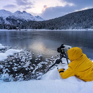 Photographer taking pictures of frozen Lej da Staz at dawn lying on the snow, Engadine