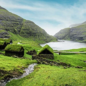 River flowing to the ocean crossing green meadows with grass-roof houses, Saksun, Streymoy Island, Faroe Islands, Denmark, Europe