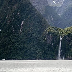 A sightseeing ship dwarfed by a tall waterfall in a fjord, South Island, New Zealand