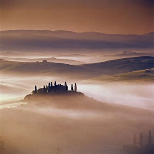 Tuscan farmhouse with cypress trees in misty landscape at sunrise