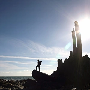 A walker stands on a boulder beside the isolated Bear Rock, a Culm pinnacle near Hartland Quay