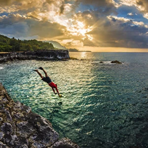 A young man dives into the calm sea on the northern rocky coastline of Kauai at sunset