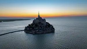 Settlement Prints: Aerial view reveals Mont St. Michel surrounded by water at sunset with cloudy sky in the background over France