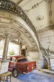 Classic red American car parked beneath ornate marble staircase inside dilapidated apartment building, Havana, Cuba