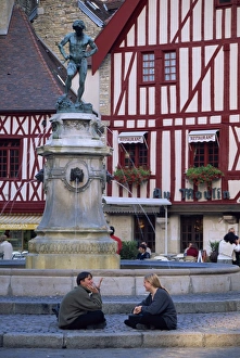 A couple sit in front of a fountain in the Place Francois Rude, with half timbered houses behind