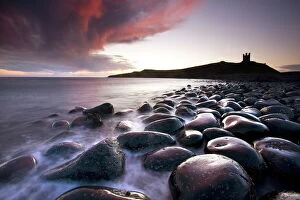 Dawn over Embleton Bay with bas alt boulders in the foreground and the ruins of Duns tanburgh Cas tle in the background