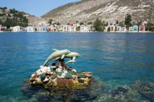 Fishermen Prints: Dolphin statues stand at harbor while boats float in Megisti Bay near colorful buildings in Greece