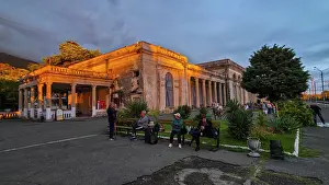 Pedestrians Prints: Gagra railway station shows people gathered near historic building during evening in Abkhazia with lights illuminating