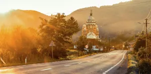 Glow Prints: Golden hour light shines on road near church in Romania with trees and mountains in background