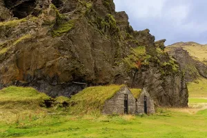 Grassland Prints: Historic turf-roofed stone houses built into cliffside near waterfall showcasing Icelandic architecture and landscape