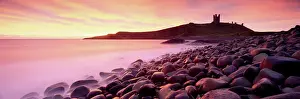 Day Break Prints: An imposing silhouette of Dunstanburgh Castle against a magnificent sky at sunrise with a beach of basalt boulders