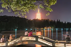 Leifeng Pagoda with visitors sitting on railings of an illuminated stone arch bridge at West Lake, Hangzhou, Zhejiang, China, Asia