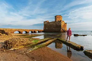 Ancient Rome Prints: Man with hat stands on top of Roman ruin facing the medieval castle of Torre Astura rising from the sea
