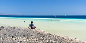 Kite Prints: Man sitting on rocks looking at the kitesurfers in the clear sea, Sotavento beach, Jandia