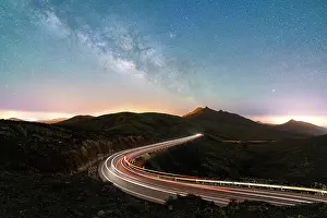 Milky Way glowing over the car trail lights on a winding desert road, Sicasumbre viewpoint, Fuerteventura, Canary Islands, Spain