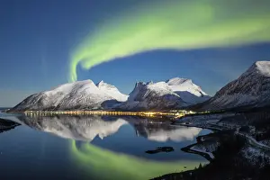 Northern lights (aurora borealis) and stars light up the snowy peaks reflected in the cold sea
