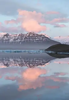 Panoramic view across the calm water of Jokulsarlon glacial lagoon towards snow-capped mountains and icebergs bathed in the last light of a winters afternoon, at the head of the Breidamerkurjokull Glacier on the edge of the Vatnajokull National Park, South Iceland, Iceland, Polar Regions