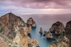 Photographer Prints: Photographer on top of cliffs surrounded by sea under the pink sky at sunrise