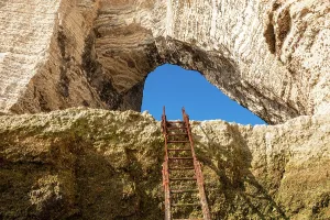 Etretat Prints: Rusty ladder leads up to natural arch with sunlight shining on chalk formations in Etretat