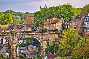 Scenic view of a historic town with a prominent stone bridge and lush greenery. Traditional architecture and a church spire add to the charm