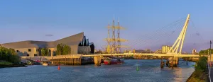 Tall Prints: Scenic view showing a bridge and a tall ship moored near modern buildings at sunset in a waterfront location