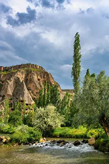Cappadocia Prints: Selime houses carved in rock near the river in Ihlara valley with trees and clouds in the sky in Cappadocia