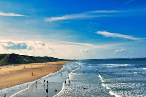 Sunny beach scene with people and rolling waves, blue sky with clouds overhead, and a hilly backdrop in Saltburn-by-the-Sea, England