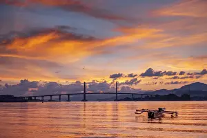 South East Asian Prints: Sunset over the harbour in Ambon city showing the suspension bridge and an outrigger boat
