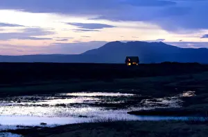 Wet Land Prints: Twilight view towards mountains with small house illuminated by interior lights