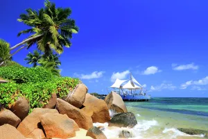 Warmth Prints: View of a beach with palm trees and rocks in front of a structure by the water at a resort in Seychelles