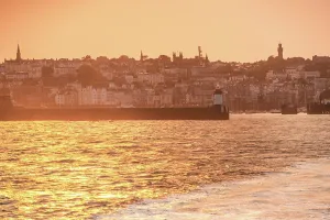 Channel Prints: Warm light shines over St. Peter Port harbor and lighthouse during sunset in Guernsey