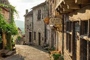Cobblestone Prints: Winding street in Saint-Cirq-Lapopie revealing stone houses and a sign for a local business in southwestern France