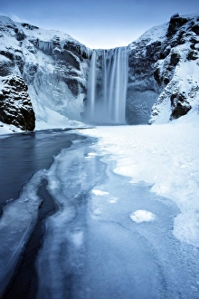 Winter view of Skogafoss waterfall, with cliffs covered in icicles and foregreound covered in snow